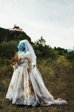 rahatsız edici bir kırsal manzara gülümseyen bir kirli ve yırtık pırtık gelin elbise giyen ve kuru çiçek, buket taşıyan bir korkunç kötü palyaço,