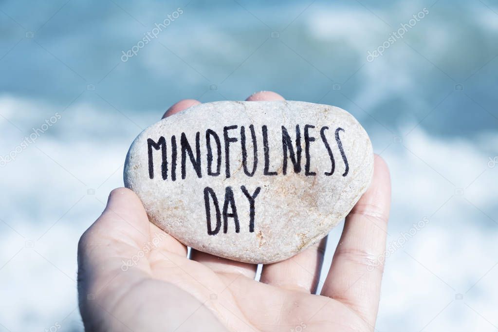 closeup of a young caucasian man, on the beach, showing a stone with the text mindfulness day written in it