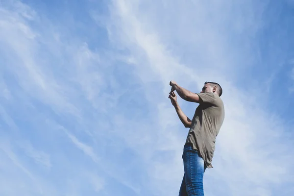 若い白人男のジーンズと グリーンの シャツ 側から見た空に対して彼のスマート フォンで写真を撮影 — ストック写真