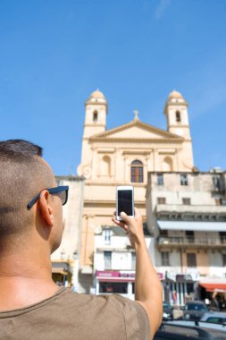 Onun smartphone ile eski şehir, Bastia, Korsika, Fransa, Saint-Jean Saint-Jean-Baptiste kilise resmini alarak güneş gözlüğü, giyen genç bir beyaz adam, closeup