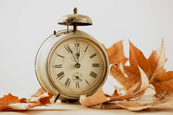 closeup of an old and rusty alarm clock surrounded by dry leaves, depicting the end of the summer time and the beginning of autumn