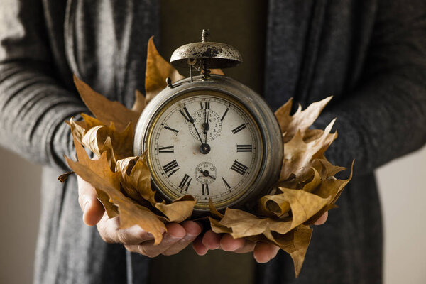 closeup of a young man with an old and rusty alarm clock surrounded by dry leaves in his hands, depicting the end of the summer time and the beginning of autumn