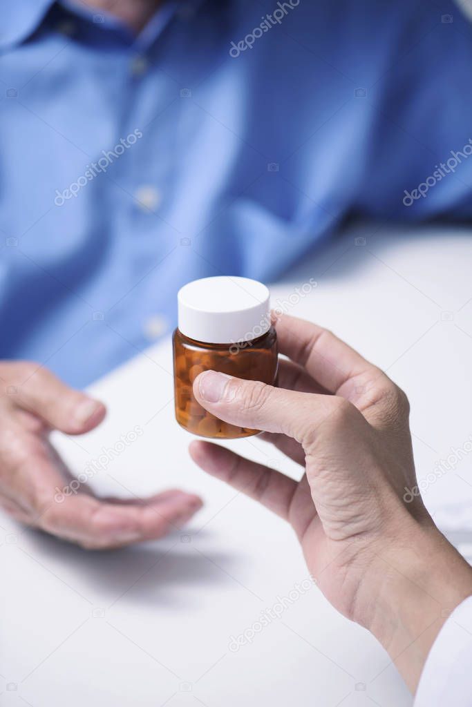 closeup of a caucasian doctor man, in a white coat, giving a bottle of pills to a senior caucasian patient man, sitting both at a doctors desk