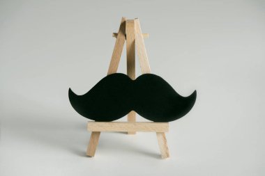 a black moustache on a wooden easel, on an on off-white background with some blank space clipart
