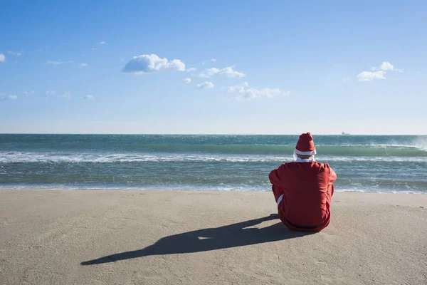 从后面看到的圣诞老人 坐在面朝大海的海滩上 望着地平线 — 图库照片