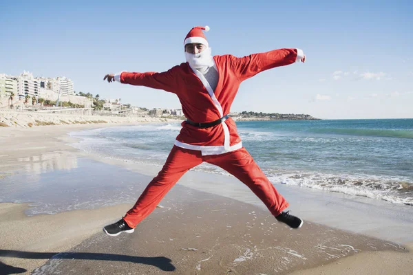 一个穿着圣诞老人西装的年轻的白种人在海滩上跳来跳去 — 图库照片