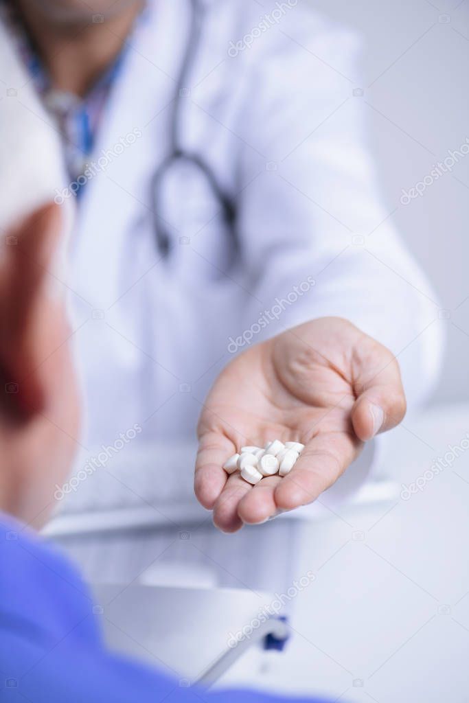 closeup of a caucasian doctor man, in a white coat, giving some pills to a senior caucasian patient man, sitting both at a doctors desk