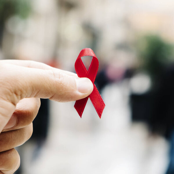 closeup of a red awareness ribbon for the fight against AIDS in the hand of a young caucasian man in a busy pedestrian street of a city