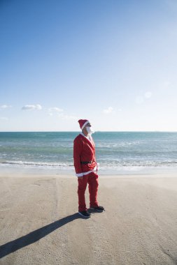 Genç adam sahilde güneşin okyanusun içinde belgili tanımlık geçmiş ile karşı karşıya duran bir Noel Baba takım elbise