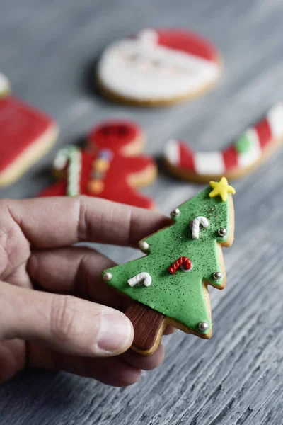 一个年轻的高加索人的特写镜头与一个手工制作的饼干形状的圣诞树男子在他的手和一堆其他不同的圣诞饼干在一个灰色的乡村木桌上 — 图库照片