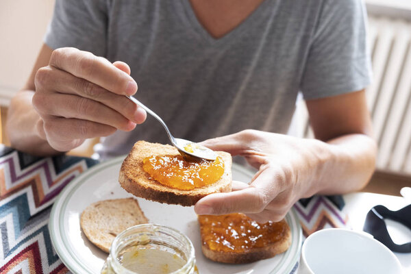 closeup of a young caucasian man, wearing a casual gray T-shirt, sitting at a set table, spreading some orange or peach jam on a toast