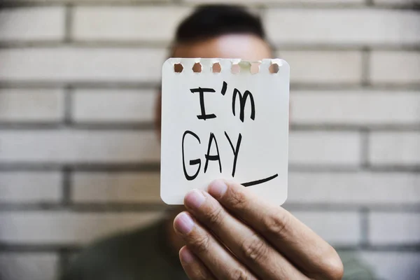 closeup of a young caucasian man holding a paper note in front of his face with the text I am gay written in it