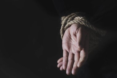 closeup of a man with his hands tied behind his back with rope, against a black background with some blank space on the left clipart