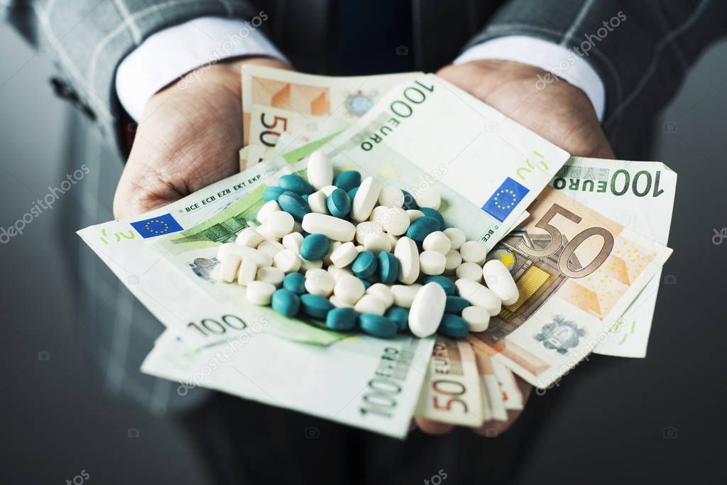 closeup of a young caucasian businessman, wearing an elegant gray suit, with a pile of euro bills and pills of different colors in his hands