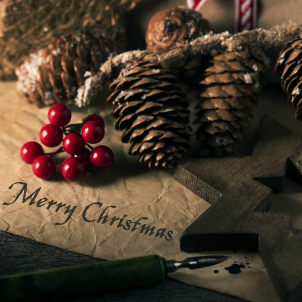 一些自然舒适的圣诞饰品的特写镜头 如松果或红色浆果 和文本快乐的圣诞蛋糕写在一张旧的纸与笔尖笔 — 图库照片