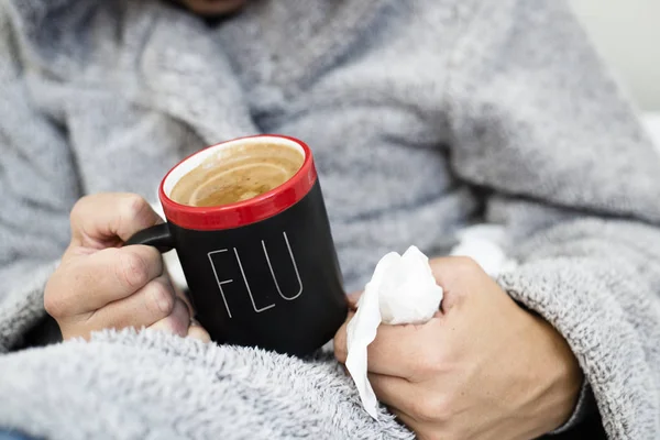 closeup of an ill young caucasian man at home wearing a fluffy house robe with a hood, warming up with a cup of hot coffee, with the word flu written in it