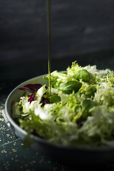 closeup of a spurt of olive oil falling on a mix of different salad leaves, like romaine lettuce, endive or arugula, on a rustic metal dish