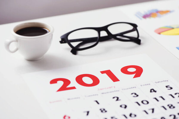 Крупный план календаря 2019 года на белом офисном столе, рядом с чашкой кофе, очками и графиками
