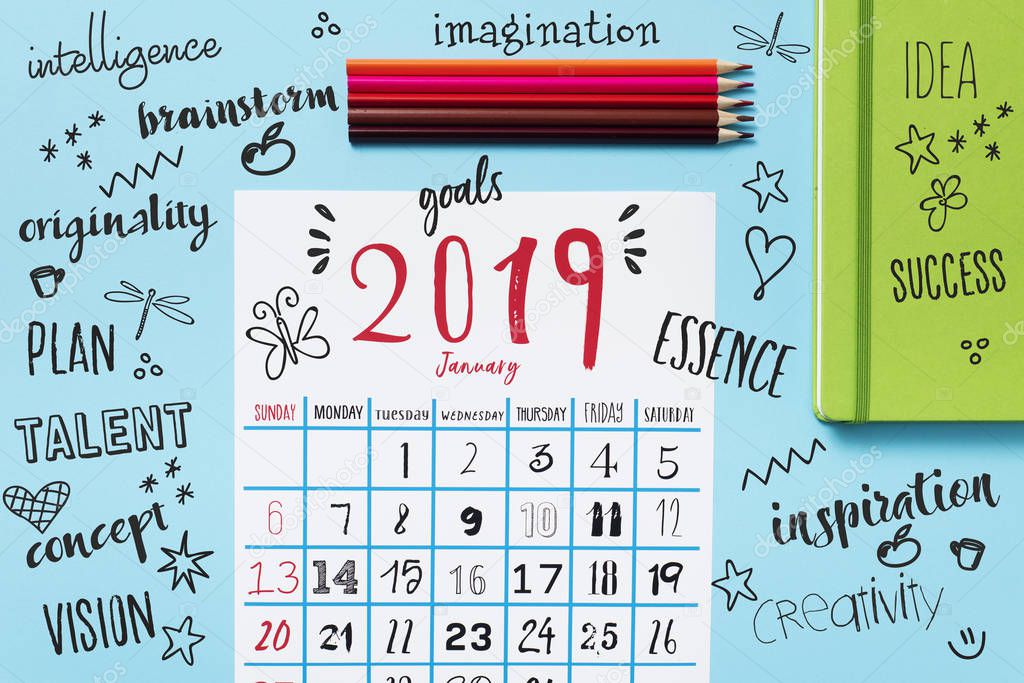 high angle view of a calendar of 2019 on a bright blue table, next to some pencil crayons of different colors and a green notepad, and some new year resolutions
