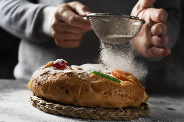 Roscon ドにいくつかの砂糖を振りかける白人男のクローズ アップ レイエス 灰色の素朴なテーブルの上に置かれた Epiphany 日食べる つのスペインの王ケーキ — ストック写真