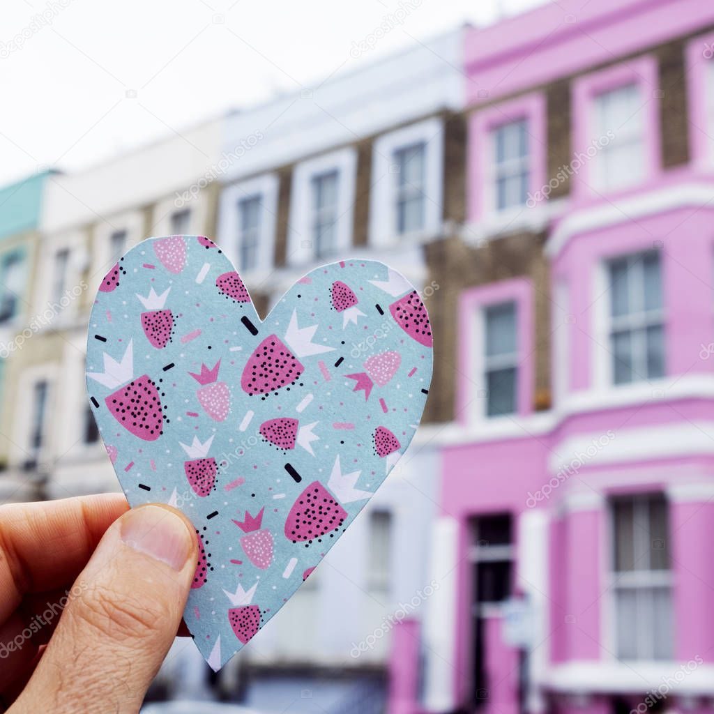 closeup of a caucasian man holding a heart cutout on a colorful paper, in front of a row of colorful houses in the popular portobello road in london, united kingdom