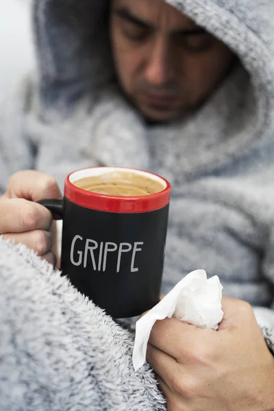 一个生病的年轻白种人在家里穿着蓬松的房子长袍与引擎盖 热身一杯热咖啡 与字抓手 流感在法国 写在它 — 图库照片