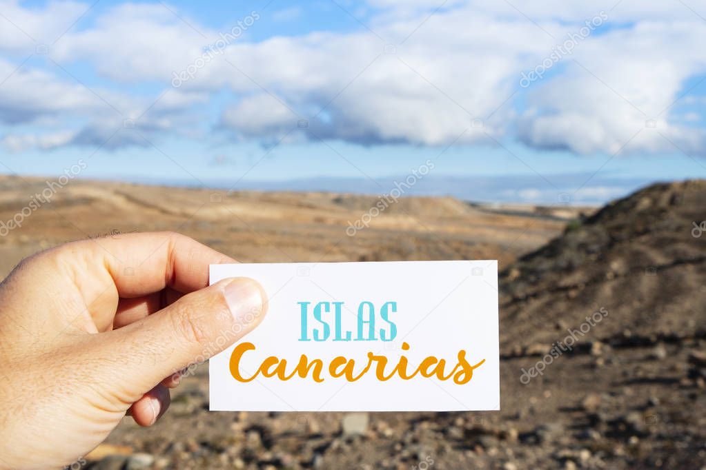 closeup of the hand of a caucasian man holding a signboard with the text Islas Canarias, Canary Islands written in Spanish, in front of a dry landscape in the Canary Islands, Spain