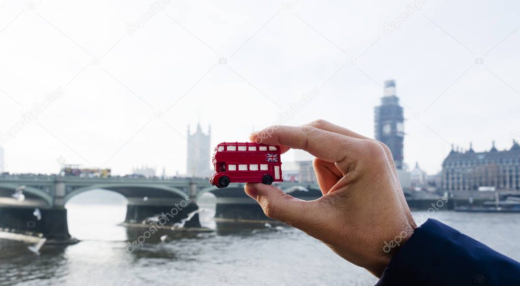 the hand of a caucasian man holding a miniature of a red double-decker bus, typical of London, United Kingdom, with the Westminster bridge and the the Palace of Westminster in the background