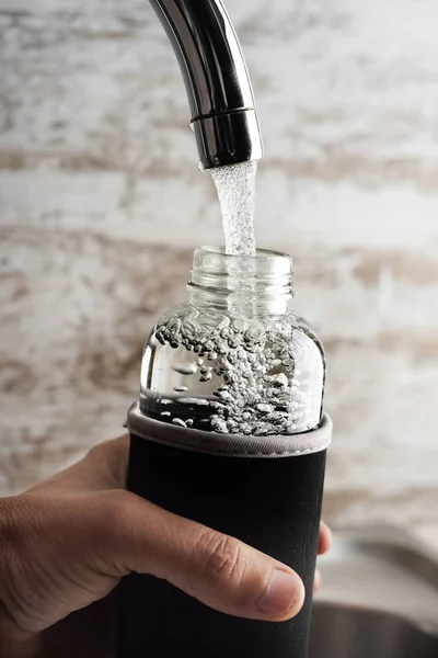 一个年轻的可可豆男子的特写镜头填充一个玻璃可重复使用的水瓶与水从一个过滤过的水站或一个家庭厨房的水龙头 — 图库照片