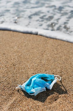 closeup of a blue used surgical mask thrown on the wet sand of the seashore of a beach clipart