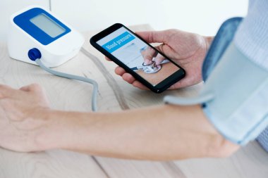 Kafkasyalı genç bir adamın yakın çekimi, kan basıncını sphygmomanometre ile ölçüyor, akıllı telefonu elinde ve ekranında kan basıncı var.
