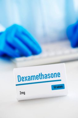 closeup of a simulated box of dexamethasone on a white table, and a man of a man wearing a white coat and blue surgical gloves in the background clipart
