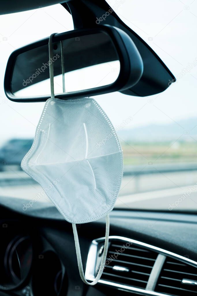 closeup of a white face mask hanging from the rearview mirror of a car on the road, while the driver is not wearing it