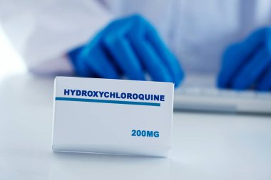 Beyaz bir masanın üzerindeki hidroklorokinin simüle edilmiş kutusunun ve arka planda beyaz önlük ve mavi ameliyat eldivenleri giyen bir adamın yakın çekimi.