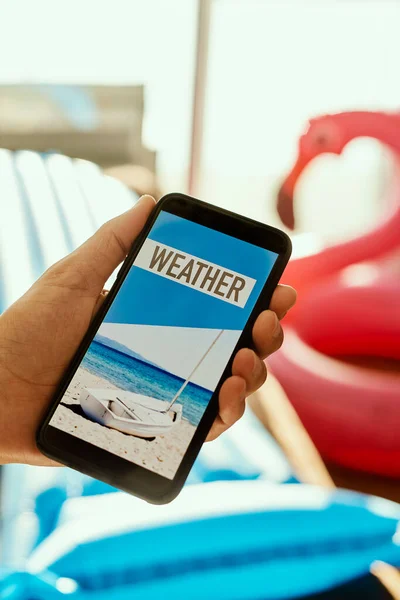 普段着の夏服を着て椅子に座りスマートフォンを手にテキストの天気を画面に映し出しピンクの水泳リングを背景に — ストック写真