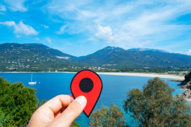 Kafkasyalı bir adamın elinde Akdeniz 'i işaret eden kırmızı bir işaretle arka planda Fransa' nın Corse eyaletinin güneybatı kıyısındaki Olmeto 'daki Plage de Baraci plajını gösteren bir el var.