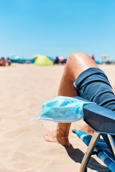一名高加索男子的特写镜头 他坐在海滩上的一张甲板椅子上 穿着蓝色短裤 头戴外科口罩挂在扶手上 稍纵即逝 — 图库照片