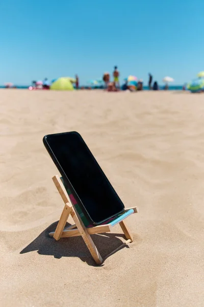 在海滩沙滩上的甲板椅子上的一部黑色智能手机 描绘了数字排毒的概念 背景上是难以辨认的人在享受和大海 — 图库照片