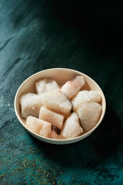 Koyu yeşil bir yüzey üzerinde pişmemiş donmuş morina balığıyla bej bir tabağın yakın çekimi.