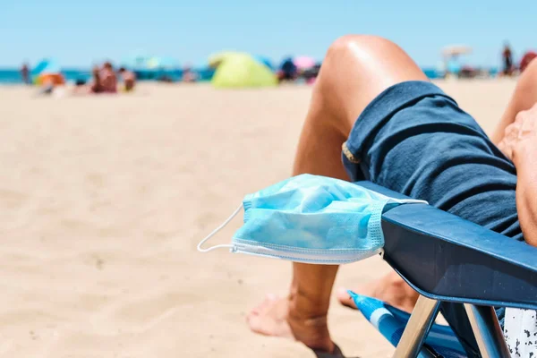 一名高加索男子的特写镜头 他坐在海滩上的一张甲板椅子上 穿着蓝色短裤 头戴外科口罩挂在扶手上 稍纵即逝 — 图库照片