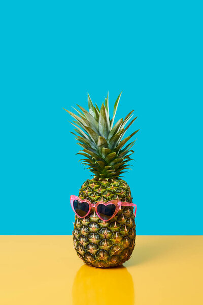 a pineapple wearing a pair of pink heart-shaped sunglasses on a yellow surface against a blue background with some blank space on top