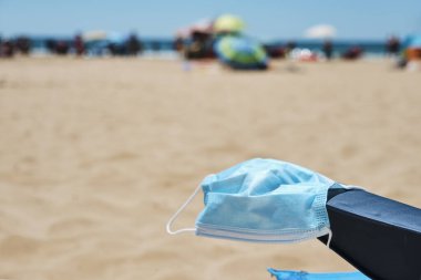closeup of a surgical mask on the armrest of a blue deck chair on the beach, with the ocean in the background clipart