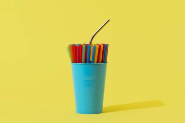 Tekrar kullanılabilen paslanmaz çelikten bir pipet mavi bir vazoda farklı renklerde pipetler içerek etraflarında boş bir alan olan sarı bir arkaplanda duruyor.