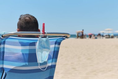 Sahilde bir iskemlede oturan beyaz bir adamın yakın çekimi, arkadan görülüyor, ve cerrahi maskesi sandalyeden sarkıyor, onu giyerken veya güneşlenirken nefes alırken