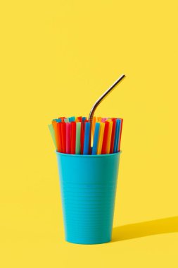 Tekrar kullanılabilen paslanmaz çelikten bir pipet. Mavi bir vazoda farklı renklerde pipetler içen tek kullanımlık plastik bir çubuk. Üzerinde boş bir alan olan sarı bir arkaplan.