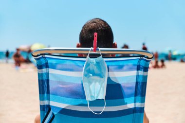 Sahilde bir iskemlede oturan beyaz bir adamın yakın çekimi, arkadan görülüyor, ve cerrahi maskesi sandalyeden sarkıyor, onu giyerken veya güneşlenirken nefes alırken