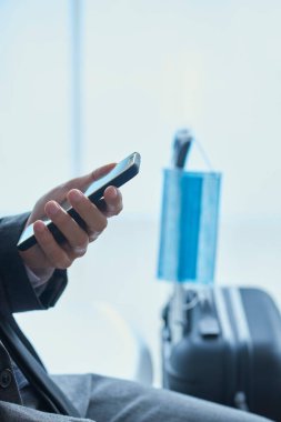 İş adamı, gri takım elbiseli, havaalanının ya da tren istasyonunun bekleme salonunda oturuyor, akıllı telefonunu kullanıyor ve arka plandaki çantasının teleskopik sapından sarkan cerrahi bir maske takıyor.