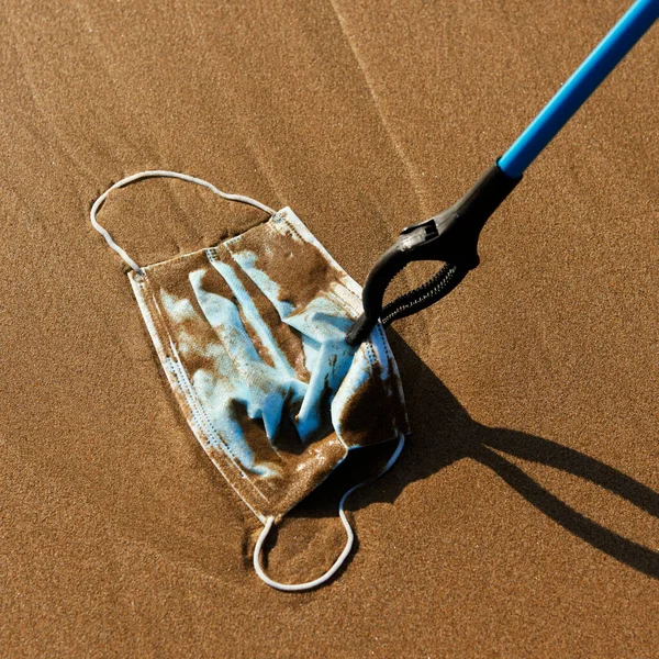 海岸のぬれた砂の上に投げ出された青の手術用マスクを回収するために手の届く範囲を使用している者の閉鎖又は海岸に海によって連れ戻された者 — ストック写真
