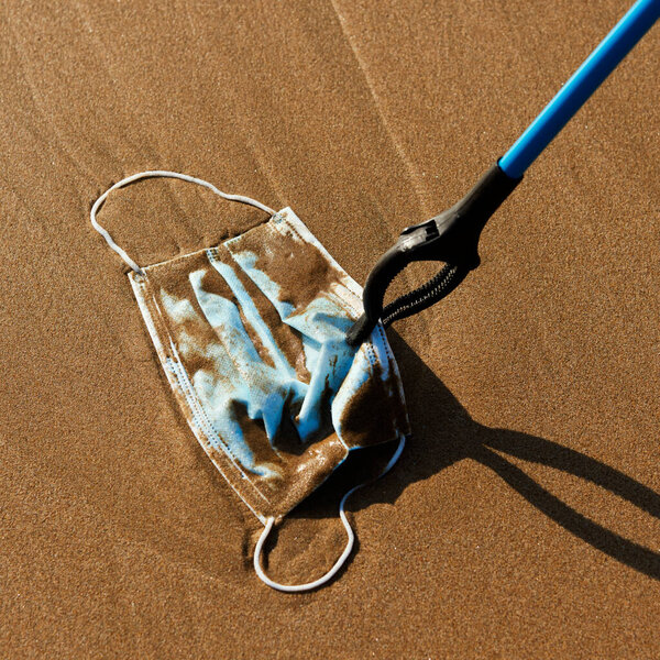 closeup of someone using a reach extend to collect a blue used surgical mask thrown on the wet sand of the seashore or brought back by the ocean to the shore