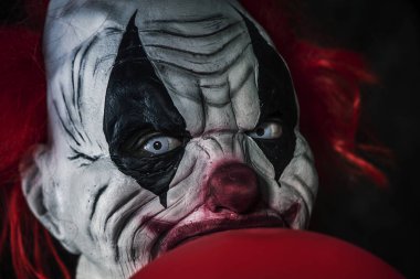 Kırmızı saçlı, beyaz gözlü, önünde kırmızı bir balonla siyah bir arka plana bakan korkunç kötü bir palyaçoya yakından bakın.